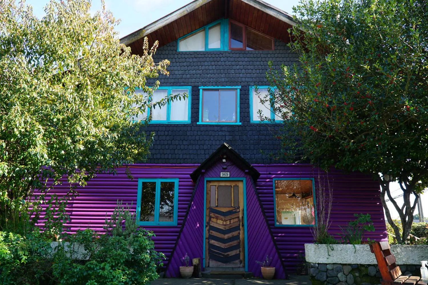 Fachada color púrpura con marcos turquesa, rodeada de árboles y luz natural que resaltan el encanto rústico y tranquilo del entorno — Hostal Micelio, Villarrica.